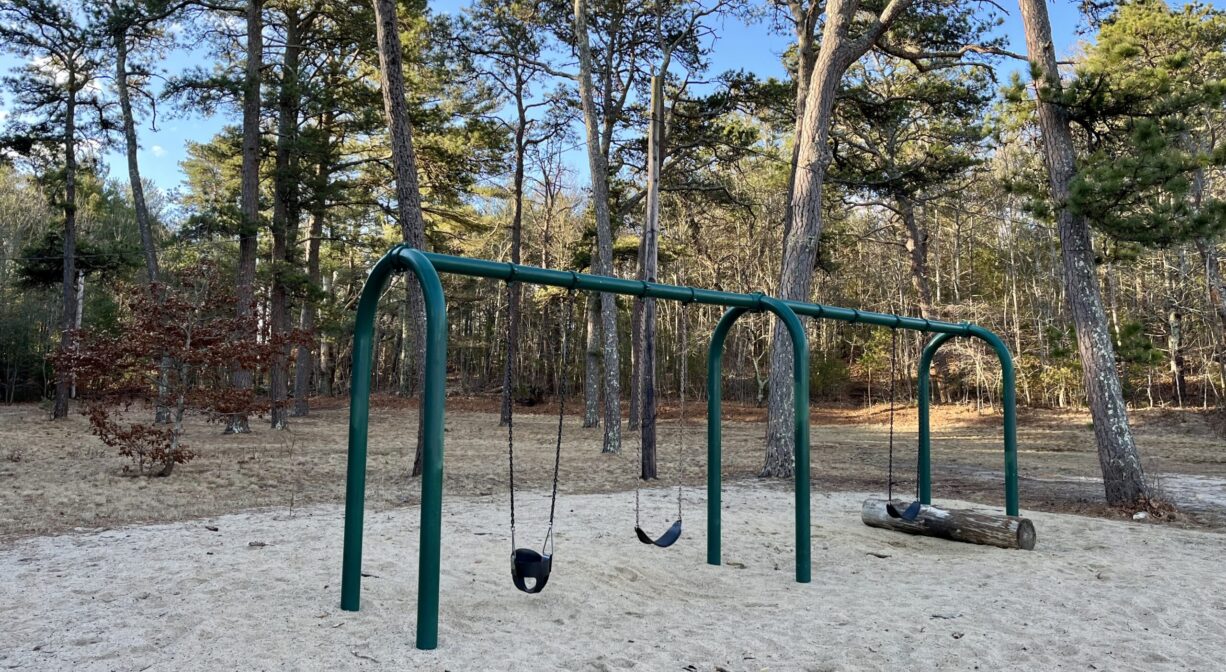 A photograph of a swing set on a sandy area with trees in the background.